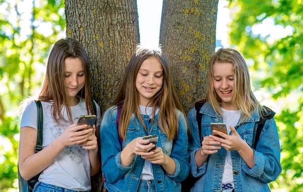 Group of three teenagers leaning against a tree looking at their mobile phones.