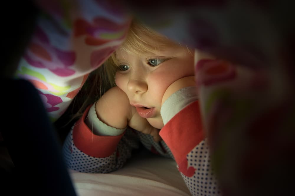 A young girl under the covers looking at a tablet screen