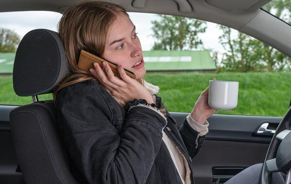 Young woman speaking on the phone and holding a coffee mug while driving with her knee.