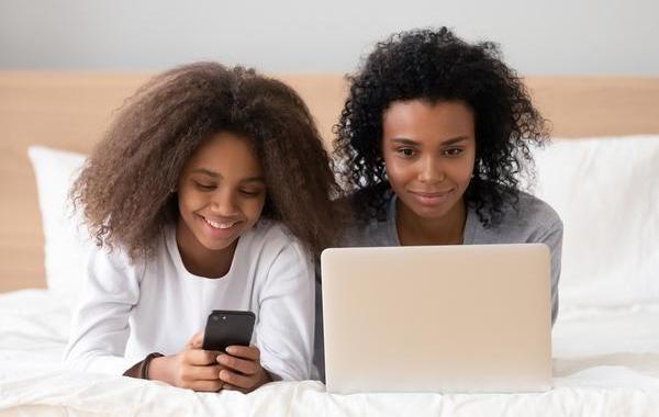 Mother and daughter laying together looking at a laptop and phone while smiling.