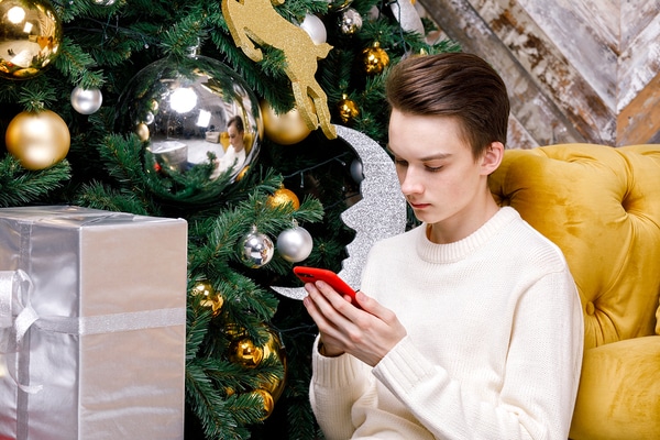Teen sitting next to a Christmas tree looking at his phone.