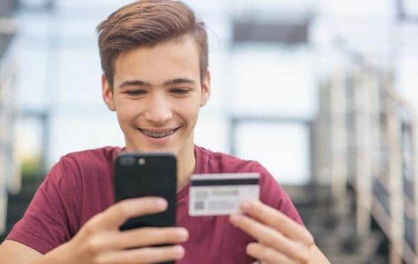 Smiling teen looking at his phone while holding a credit card.