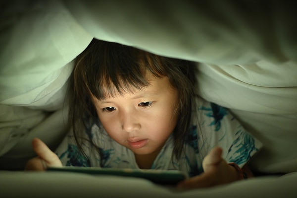 Girl lying in bed with covers over her head looking at a tablet.