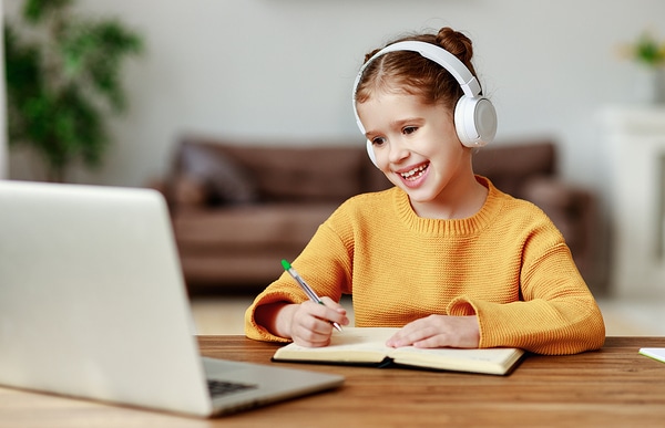 Smiling young teen girl wearing headphones, while looking at her computer screen and writing in a book.