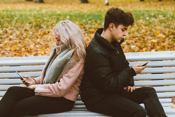 Two people sitting on a park bench looking at their phones.
